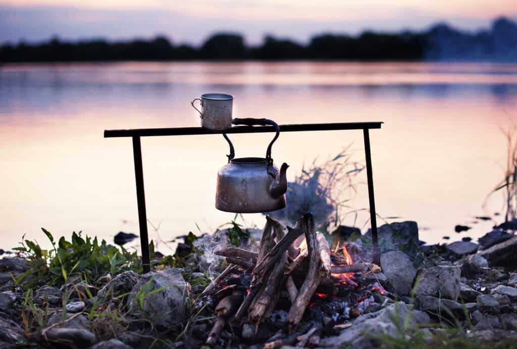 Bivouac sur la rivière au coucher du soleil, avec une bouilloire suspendue au-dessus du feu pour le thé et le café.