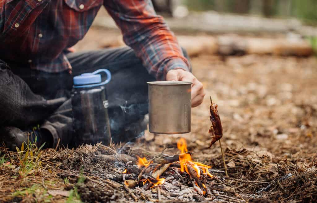 homme voyageur mains tenant une tasse avec de l'eau près du feu en plein air